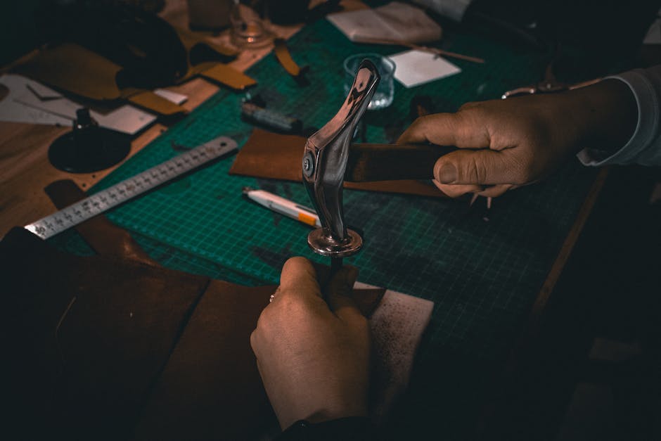 Hands crafting leather with tools on a workbench under warm lighting, showcasing artisanal skill.